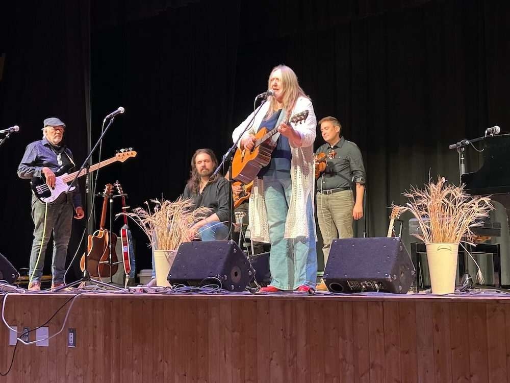 A musical performance on stage with a group of musicians playing various instruments, surrounded by decorative plants and instruments